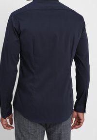 Camisa de manga larga azul marino con cuello clásico, puños abotonados y una textura suave. Presenta un dobladillo curvo y sin patrones visibles.