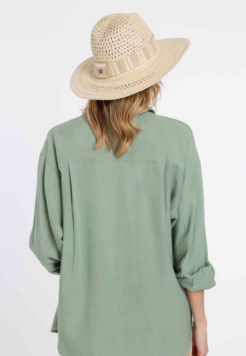 Femme aux cheveux châtain clair portant une chemise verte et un chapeau de soleil tissé beige, vue de dos sur un fond uni.