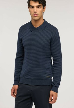 Giovane uomo che indossa un maglione blu scuro a maniche lunghe con collo e pantaloni scuri su uno sfondo chiaro e semplice.