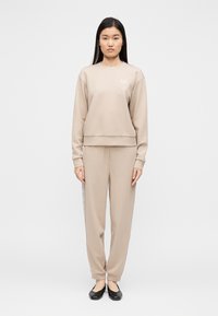 Μπεζ φούτερ και αντίστοιχο παντελόνι φόρμας από απαλό ύφασμα. Το φούτερ έχει στρογγυλή λαιμόκοψη και λογότυπο, ενώ το παντελόνι διαθέτει λεπτομέρεια πλαϊνής ρίγας.