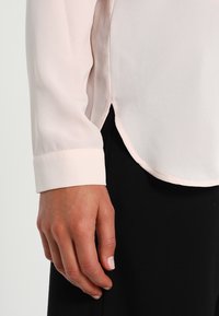Blusa rosa chiaro realizzata in tessuto morbido, con orlo arrotondato e maniche a giro. Il design presenta sottili spacchi laterali per facilitare i movimenti.