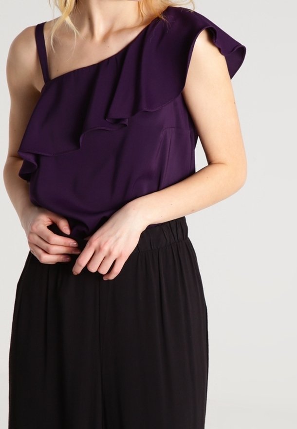 Fialový top na jedno rameno s vrstvenými volány, vyrobený z hladkého materiálu. V kombinaci s volnými černými kalhotami, které vytvářejí uvolněnou siluetu.
