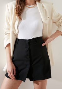 Černá sukně s knoflíkovou légou vpředu a střihaným střihem, doplněná žebrovaným bílým topem a krémovým sako, s hladkou texturou a čistými liniemi.
