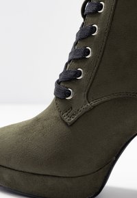 Bottine en suède vert olive avec un bout pointu et un talon aiguille. Présente des lacets noirs passés à travers des œillets en argent. Texture lisse.