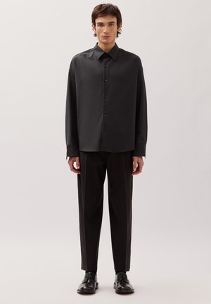 Giovane uomo in piedi, indossa una camicia nera larga con bottoni, pantaloni neri su misura e scarpe eleganti in pelle nera, su uno sfondo chiaro e uniforme.