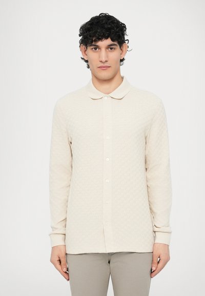 Polo à manches longues beige clair avec un motif texturé, col à revers et boutonnière sur le devant. Poignets finis avec un détail côtelé.