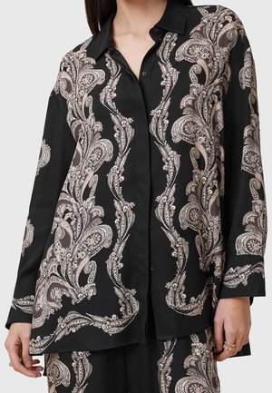 Kobieta ubrana w czarną koszulę na guziki z długim rękawem w beżowy wzór paisley oraz dopasowane spodnie, stojąca z rękami swobodnie opuszczonymi.