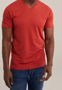 Röd kortärmad t-shirt med v-ringning, tillverkad av ett mjukt tyg. I kombination med blå jeans som visar en figursydd silhuett.