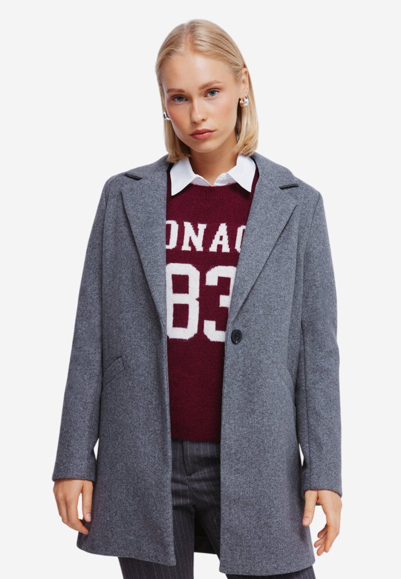 Grå uldfrakke med en struktureret design, med nothals og enkeltknaplukning, lagd over en mørkerød sweater med hvid skrift.