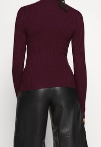 Pull en maille côtelée couleur bordeaux avec un col montant et des manches longues, associé à un pantalon en cuir noir taille haute.