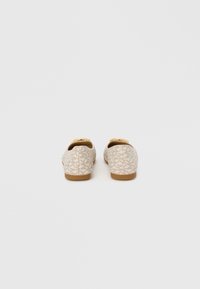 Chaussures beiges avec un motif géométrique abstrait. Semelle plate à texture marron clair. Vue arrière révélant un talon arrondi et un design simple.