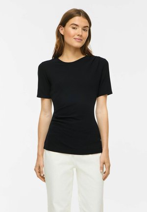 Femme aux cheveux bruns portant un haut noir ajusté à manches courtes et un pantalon blanc, debout devant un fond blanc uni.