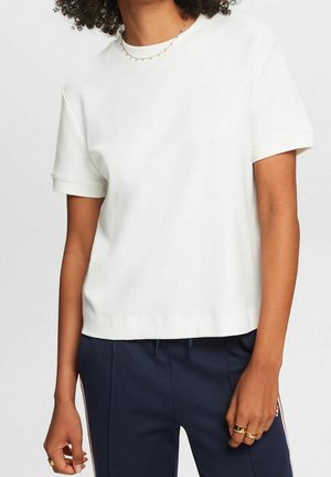 Person trägt ein schlichtes weißes Kurzarmhemd und dunkelblaue Hose mit Kordelzug, zeigt Oberkörper und Arme, steht vor einem schlichten Hintergrund.