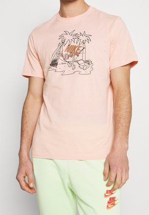 Man draagt een lichtroze Nike T-shirt met een ontwerp van een palmboom en tropische motieven, gecombineerd met lichtgroene shorts met drie Nike-logo's.