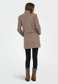 Langer, strukturierter, hellbrauner Mantel mit hohem Kragen, getragen über schwarzen Hosen. Verfügt über einen geraden Schnitt und minimalistische Designmerkmale.