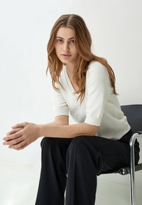 Suéter blanco de manga corta con escote en V, combinado con pantalones de pierna ancha negros, sentado en una silla metálica contra un fondo neutro.