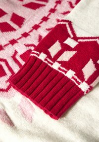 Rot und rosa gemusterter Pullover mit einem gerippten roten Bündchen, dekoriert mit geometrischen Formen und kontrastierenden weißen Akzenten auf einem sanften rosa Hintergrund.
