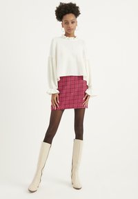Blusa bianca corta con scollatura a volant e maniche a sbuffo abbinata a una mini gonna a quadri rossi. La modella indossa stivali beige al ginocchio.