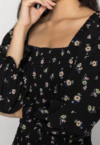 Czarna bluzka w kwiaty z kwadratowym dekoltem, marszczonym przodem, długimi bufkami oraz detalem w postaci wiązania w talii. Posiada wielokolorowe wzory kwiatowe.