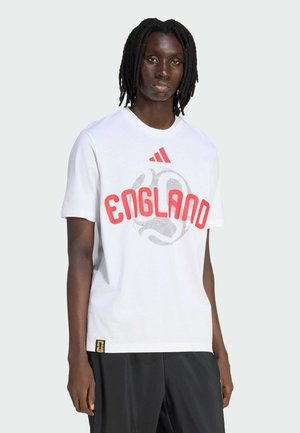 Persona con rastas que lleva una camiseta blanca con la palabra "ENGLAND" y un gráfico estilizado de un balón de fútbol en rojo y gris, combinada con pantalones negros.