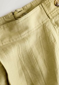 Gros plan sur la ceinture d'un pantalon beige avec un bouton et une fermeture éclair, montrant la texture du tissu et les détails des coutures.