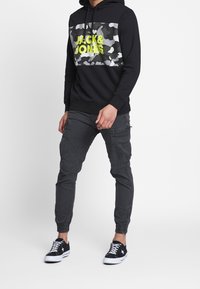 Černá mikina s šedým maskáčovým panelem, která má na sobě nápis "JACK & JONES" v neonově žluté barvě. V kombinaci s tmavě šedými cargo kalhotami. Černé sneakers.