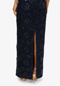 Abito blu navy con design in pizzo floreale e paillettes, spacco laterale e tacchi beige visibili. La texture del tessuto liscia esalta l'eleganza.