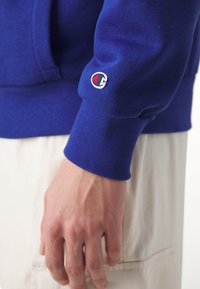 Niebieska bluza z ribb długimi rękawami; na rękawie ma małe haftowane logo. Materiał wydaje się miękki i teksturowany.