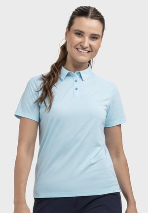 Lächelnde Frau mit braunen Haaren im Pferdeschwanz, trägt ein hellblaues kurzärmeliges Polohemd und dunkle Hose, steht vor einem schlichten Hintergrund.