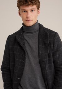 Svart och grå tartanmönstrad kappa med notch-krage och knappstängning, lager på lager över en mörkgrå polotröja. Texturerat tyg, skräddarsydd passform.