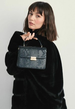 Czarna, teksturowana torebka z faux skóry z wzorem przypominającym skórę gada, z górnym uchwytem i złotymi akcesoriami. Noszona przez osobę w czarnym płaszczu.