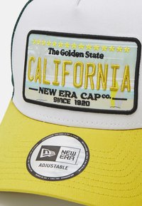 Gorra ajustable con frente blanca, visera amarilla y acentos verdes. Presenta "CALIFORNIA" bordado con estrellas y detalles de marca.
