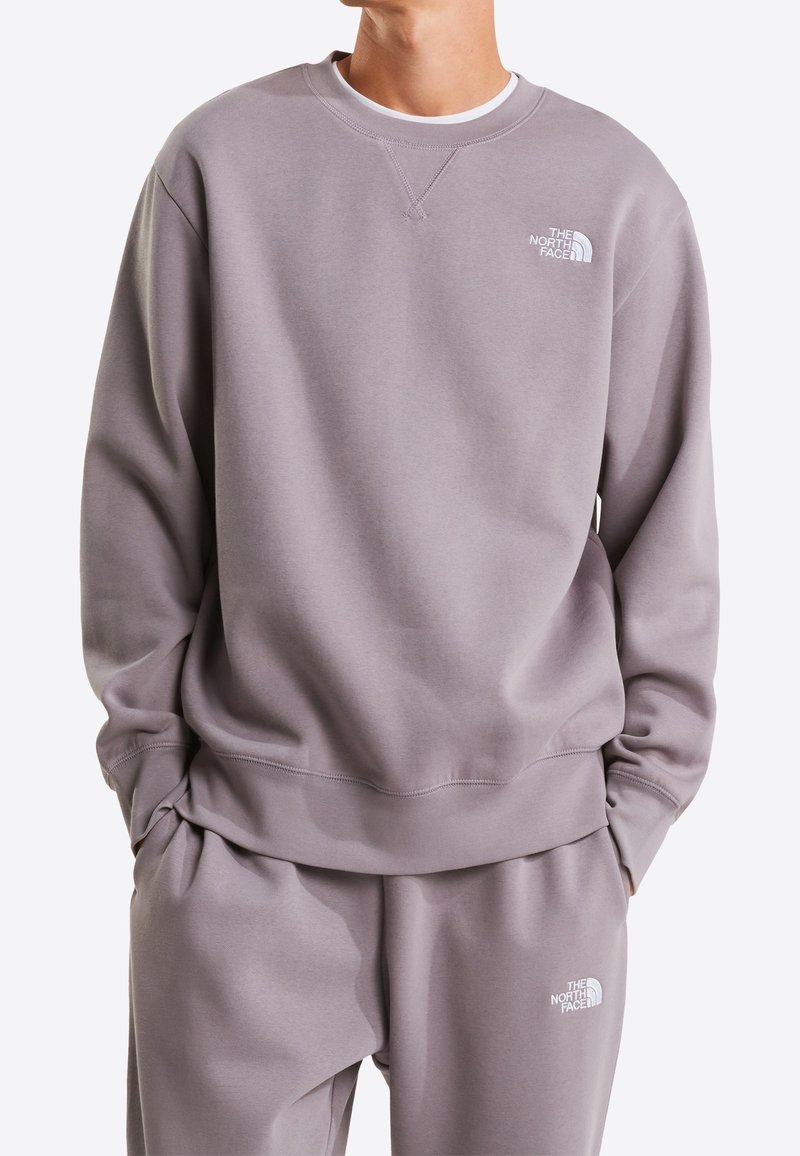Personne portant un sweat-shirt et un pantalon The North Face violet clair assortis, mains dans les poches, fond uni.