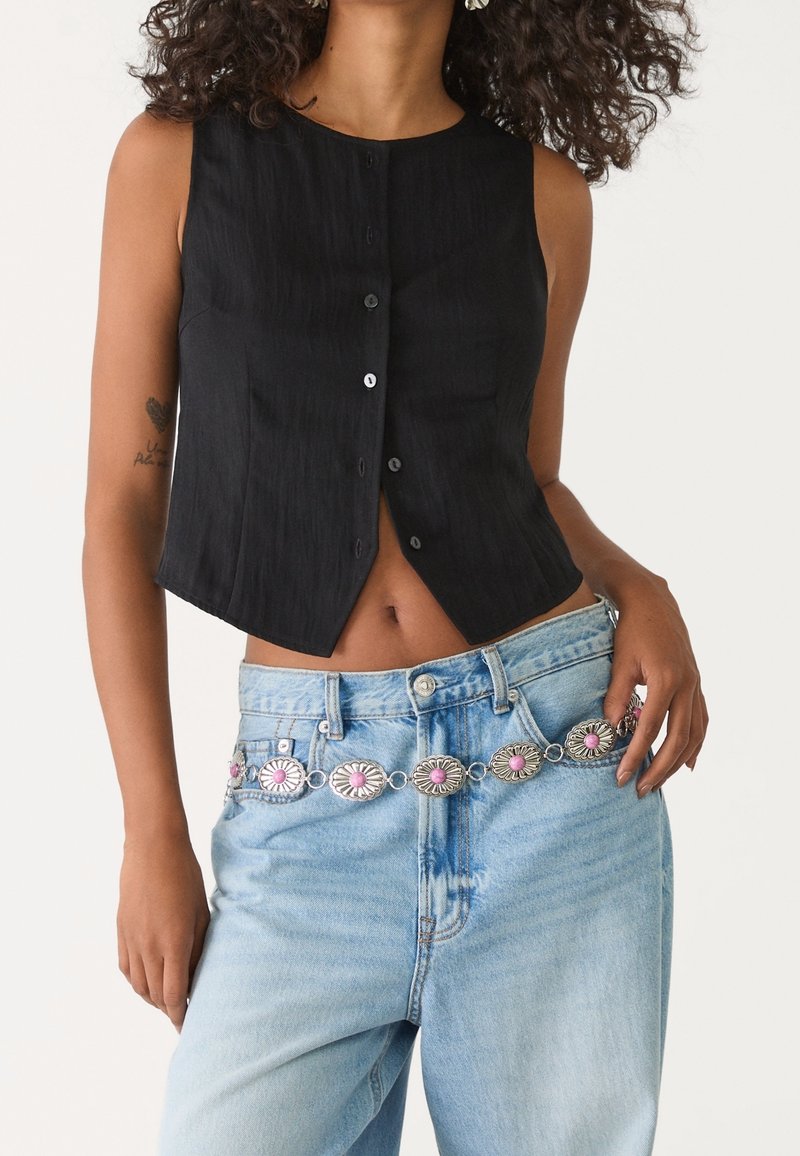 Femme portant un crop top noir sans manches boutonné, un jean bleu clair et une ceinture argentée avec des détails floraux roses.