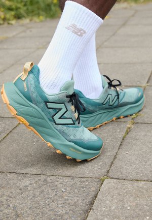 Teal and green running shoes feature a mesh upper, black logo, and a rugged, orange outsole with Vibram detailing. Worn with white socks.