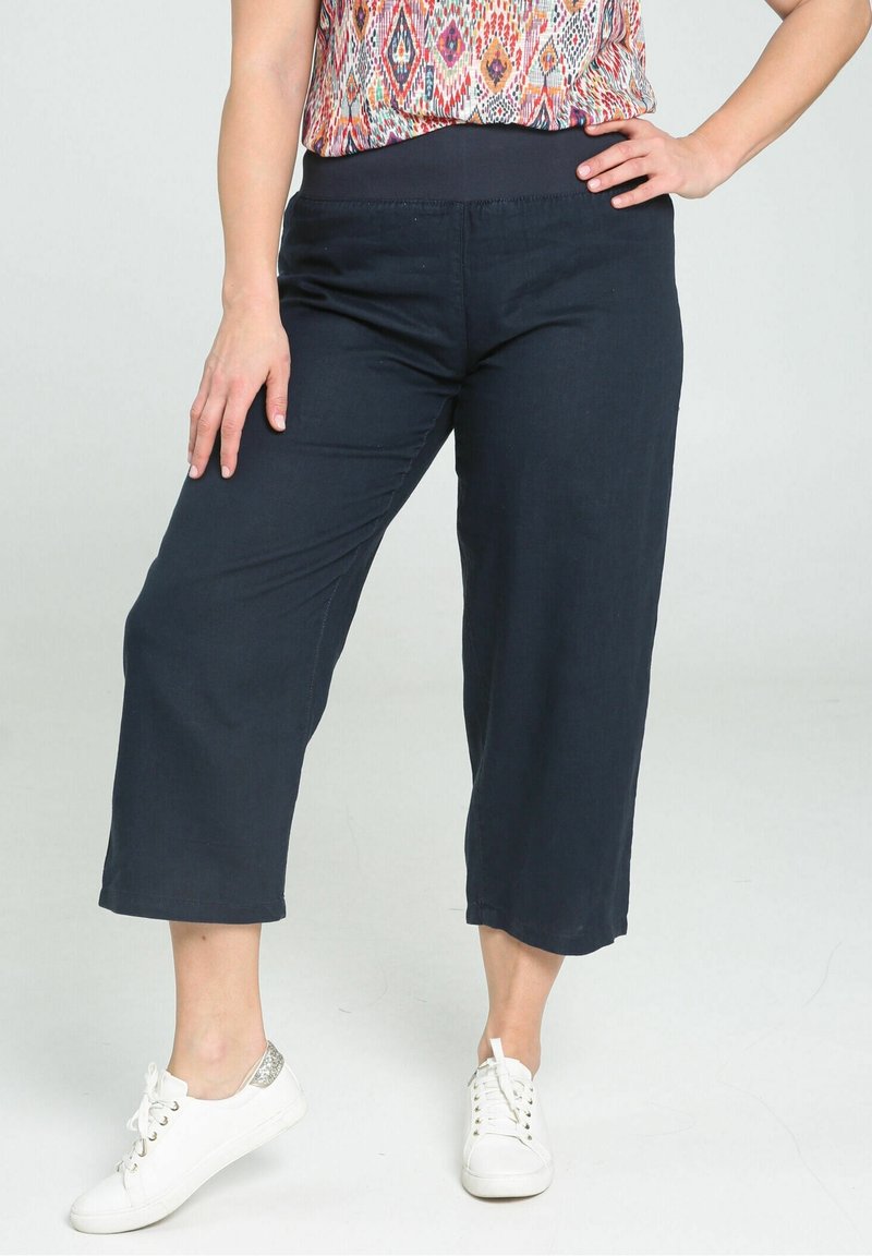 Femme portant un pantalon noir large et court, un haut coloré à motifs et des baskets blanches, debout avec une main sur la hanche.