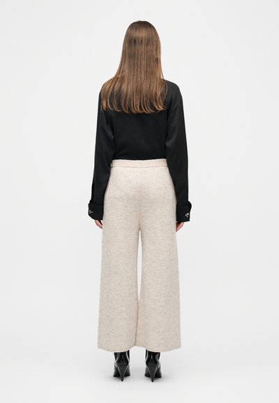 Γυναίκα που στέκεται με την πλάτη γυρισμένη, φοράει μαύρη μπλούζα με μακριά μανίκια, μπεζ παντελόνι φαρδύ στο πόδι και μαύρα μποτάκια με τακούνι, σε λευκό φόντο.