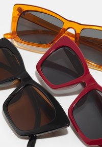 Štiri pare sončnih očal: oranžne in rdeče pravokotne oblike, črne mačje oči, vse s temnimi lečami in sijajnimi plastičnimi okvirji.