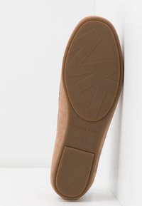 Semelle d’une chaussure plate en daim beige avec une semelle en caoutchouc texturée, présentant un grand logo MK et le marquage « Michael Kors ».