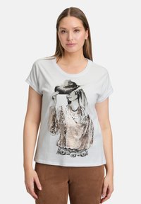 T-shirt blanc à manches courtes avec un graphique représentant une femme portant un chapeau gris et des colliers superposés, avec des détails métalliques sur le t-shirt.