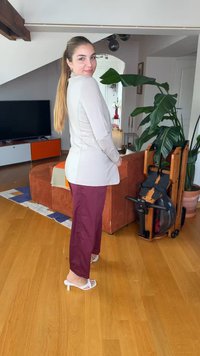 Γυναίκα με μακριά μαλλιά, φοράει λευκό μπουφάν και μπορντό παντελόνια, στέκεται χαμογελαστή στο σαλόνι με πορτοκαλί καναπέ, μεγάλο φυτό και όργανο κωπηλασίας.