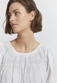 Blouse blanche avec un col rond, devant plissé et accents de broderie florale. Le tissu est léger avec une finition légèrement texturée.