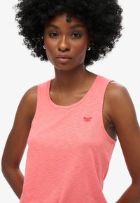 Top sin mangas de color rosa claro, con una ligera textura jaspeada y un pequeño logo rojo de "Superdry" en la parte frontal izquierda.