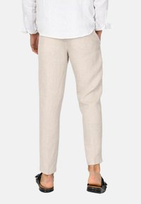 Pantaloni in lino beige con vestibilità affusolata e tasche laterali, indossati con sandali neri a scivolo. Il tessuto presenta una texture sottile.