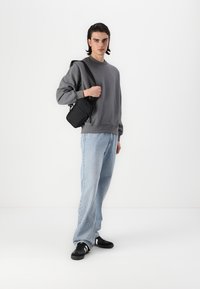 Felpa grigia con maniche lunghe, jeans blu chiaro, sneakers nere e una piccola borsa a tracolla nera, su uno sfondo bianco semplice.