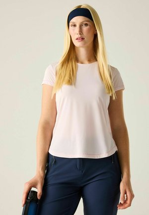 Mujer rubia con una diadema negra, camiseta rosa pálido, pantalones azul marino, sosteniendo una botella de agua azul oscuro sobre un fondo claro.