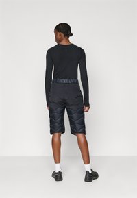 Camisa preta de manga longa combinada com calções escuros acolchoados até aos joelhos, apresentando um cós brilhante e padrões texturizados no tecido.