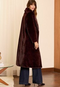 Cappotto in pelliccia sintetica bordeaux con ampio colletto, vestibilità oversize e maniche lunghe. Abbinato a pantaloni a gamba larga scuri e scarpe con tacco.