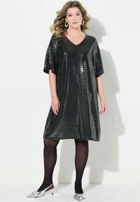 Schwarzes Pailletten-Kleid mit V-Ausschnitt und kurzen Ärmeln, das einen lockeren Schnitt und eine glatte, glänzende Textur aufweist. Kombiniert mit silbernen Schuhen.
