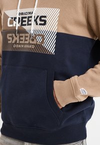 Felpa con design color block in tonalità beige, blu navy e dettagli a righe. Realizzata in tessuto morbido, include una tasca frontale e cappuccio con coulisse.